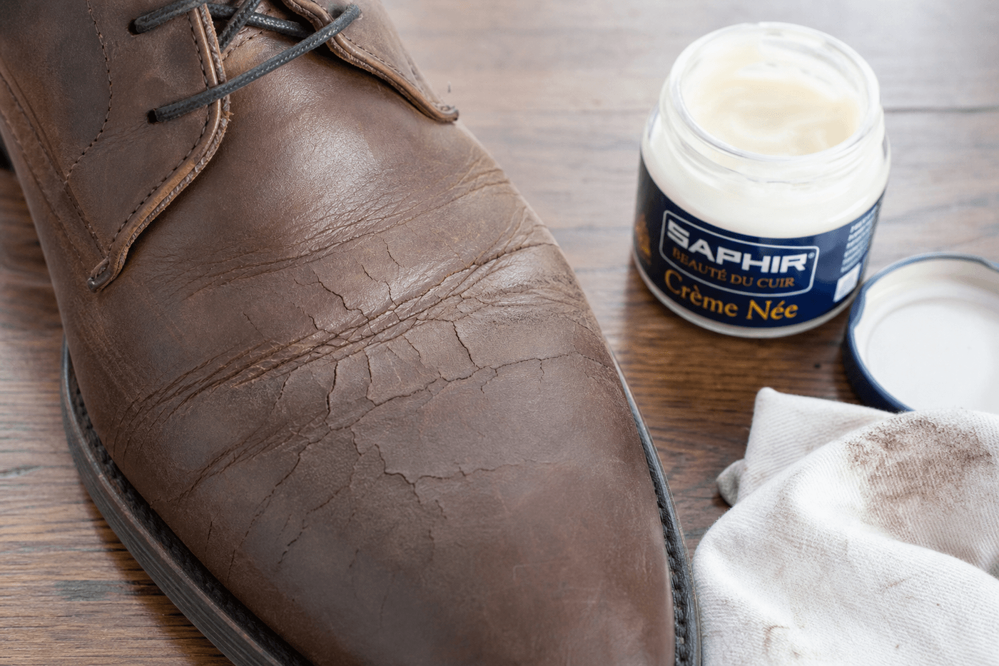 Chaussure en cuir marron avec des craquelures visibles au niveau du pli de marche, posée sur une table avec un chiffon et une crème nourrissante