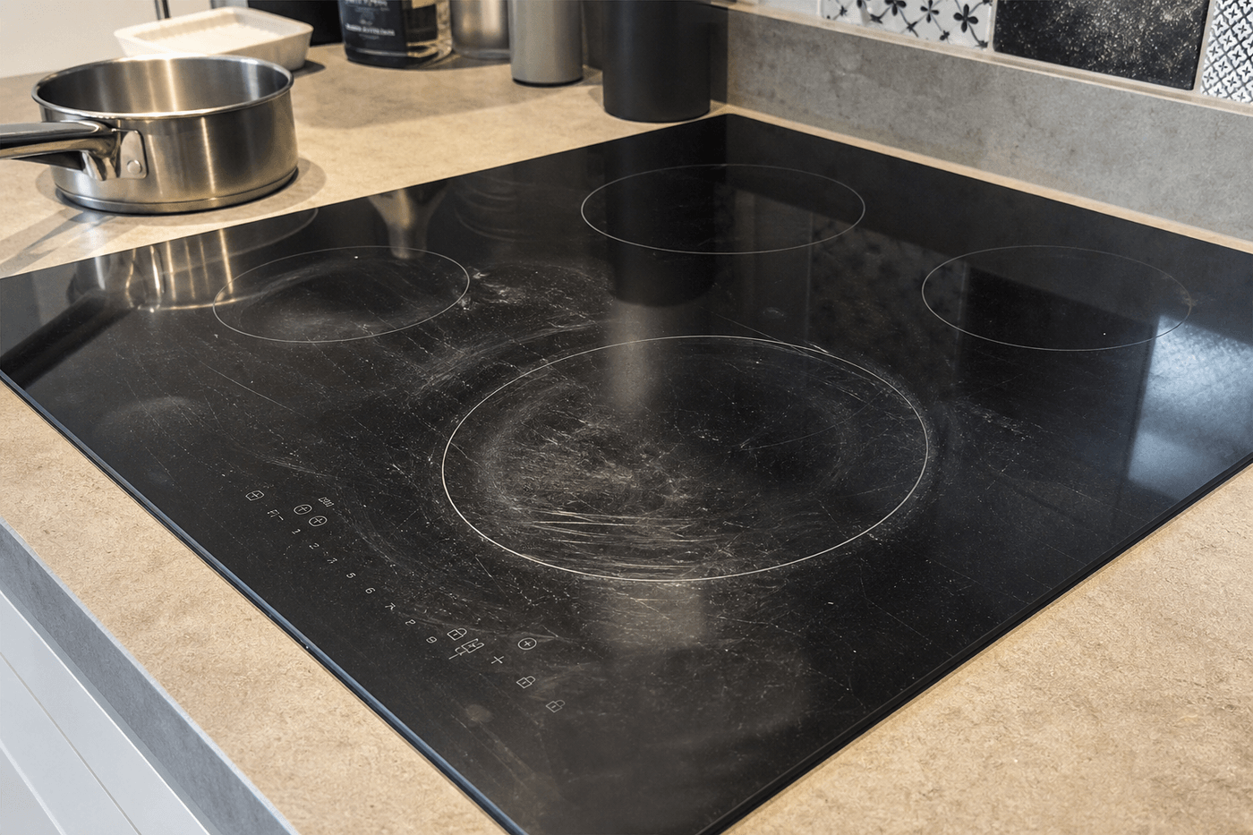 Plaque de cuisson à induction noire avec des traces blanches et des micro-rayures visibles à la lumière, casserole posée à côté