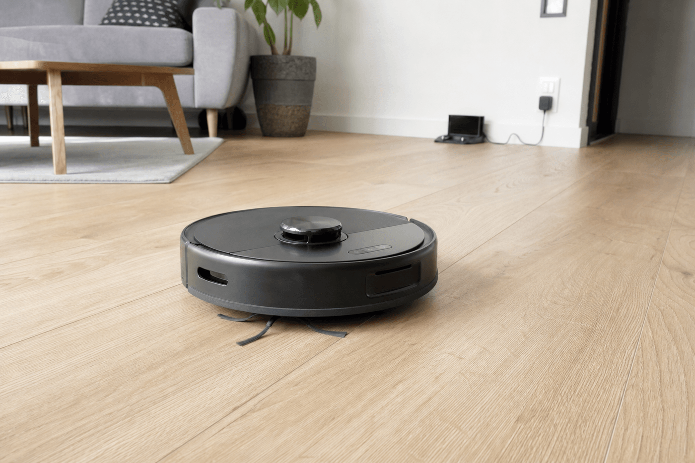Robot aspirateur arrêté au milieu d'un salon, loin de sa base de charge visible en arrière-plan contre un mur