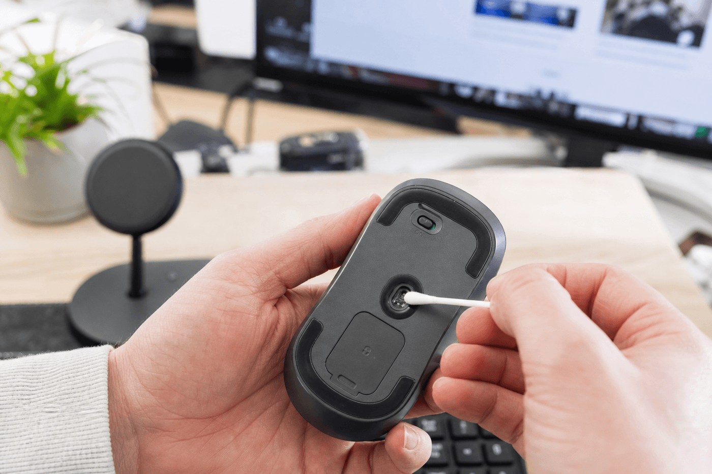 Personne nettoyant le capteur optique sous une souris d'ordinateur retournée, bureau lumineux avec écran en arrière-plan