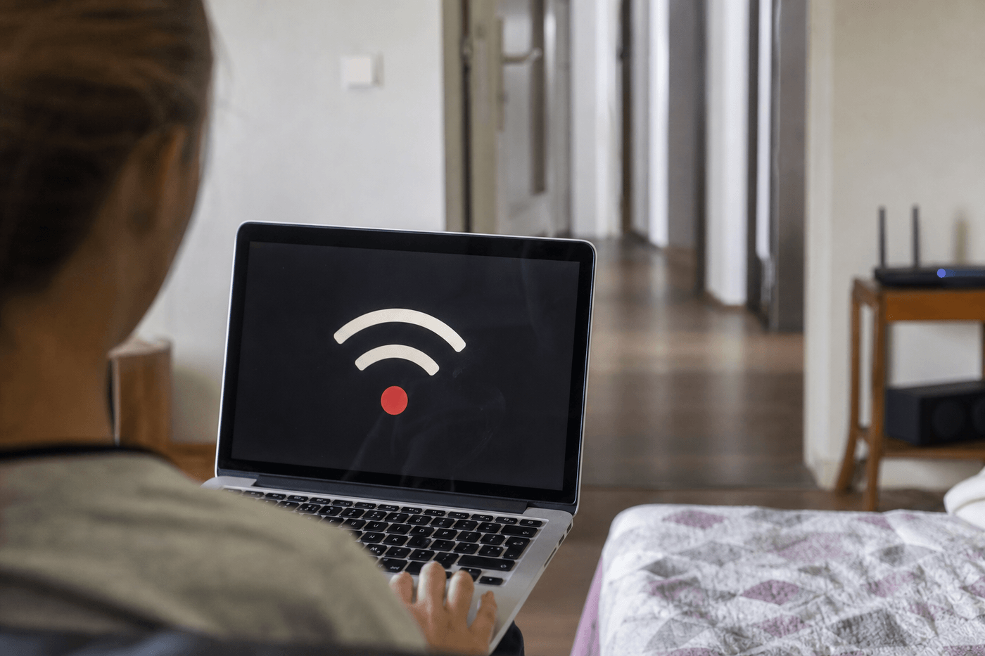 Personne dans une chambre éloignée regardant un ordinateur portable affichant un signal Wi-Fi faible, box internet au loin